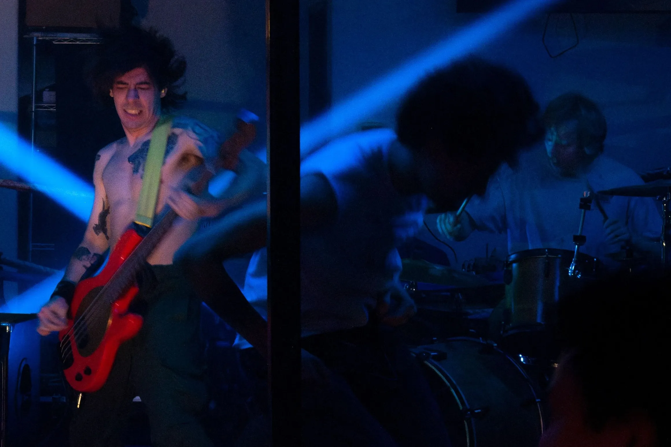 A guitarist and drummer in a tight room cut through by blue light